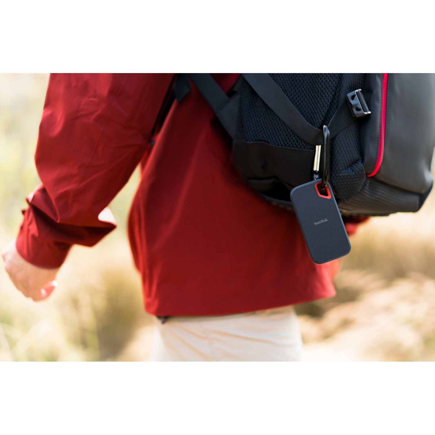 Una persona con una chaqueta roja sostiene un disco duro portátil negro sujeto a una mochila.