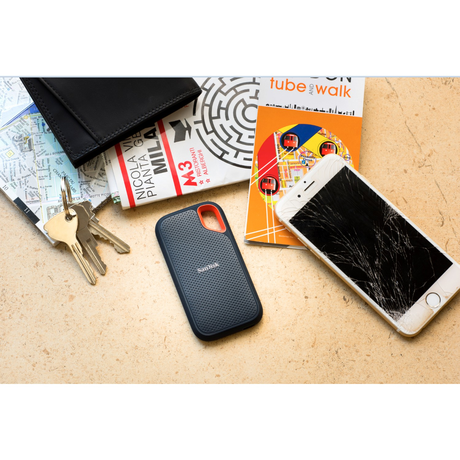 Un teléfono roto, cartera, llaves, cuaderno y disco duro externo SanDisk sobre superficie beige.