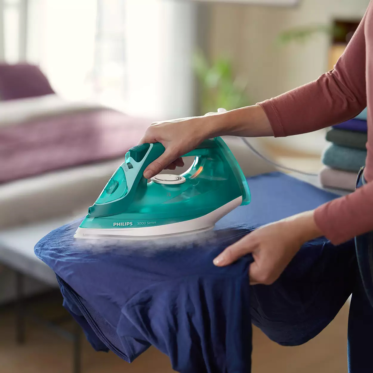 Un fer à repasser turquoise fume une chemise bleue sur une planche. Un lit et des serviettes en arrière-plan.
