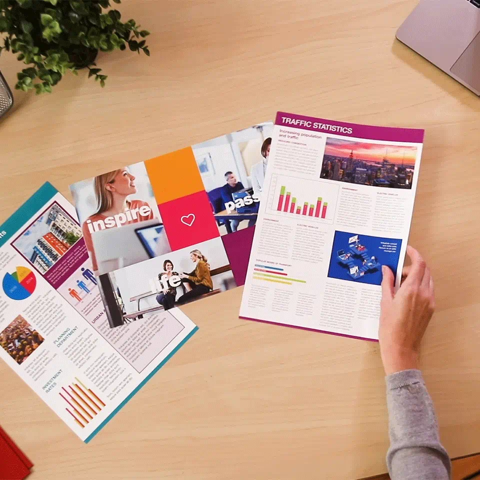 Person hält Papiere auf einem Holztisch. Papiere zeigen Bilder, Diagramme und Text. Eine Pflanze und ein Laptop im Hintergrund.