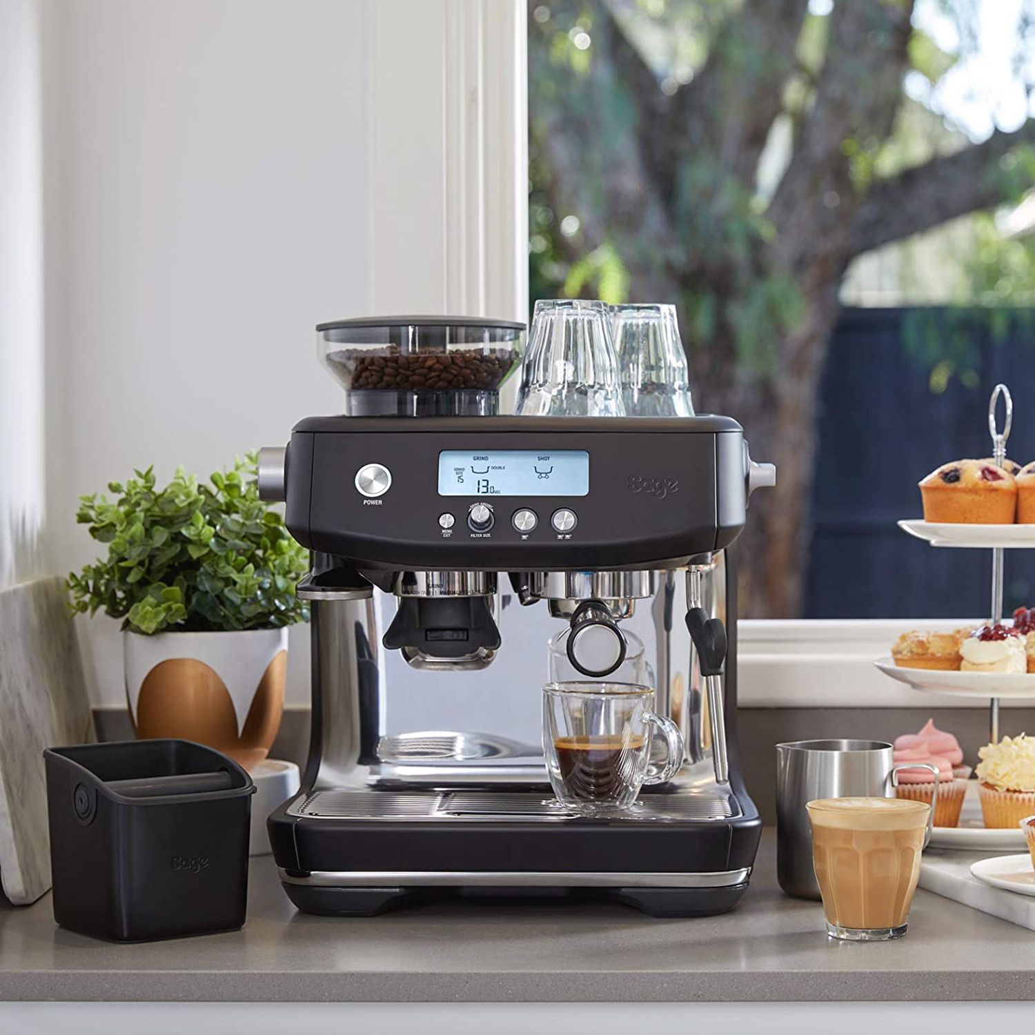 Macchina per espresso sul bancone, bicchiere di caffè, macinacaffè con chicchi, muffin e pianta.