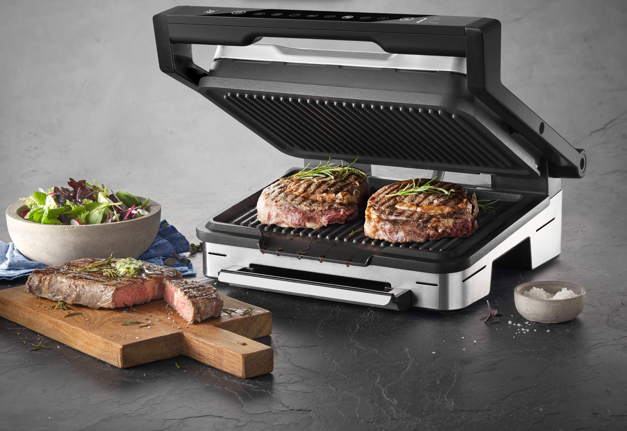 Ein offener schwarzer Grill mit zwei Steaks, einer Salatschüssel und einem Schneidebrett. Grauer Hintergrund.