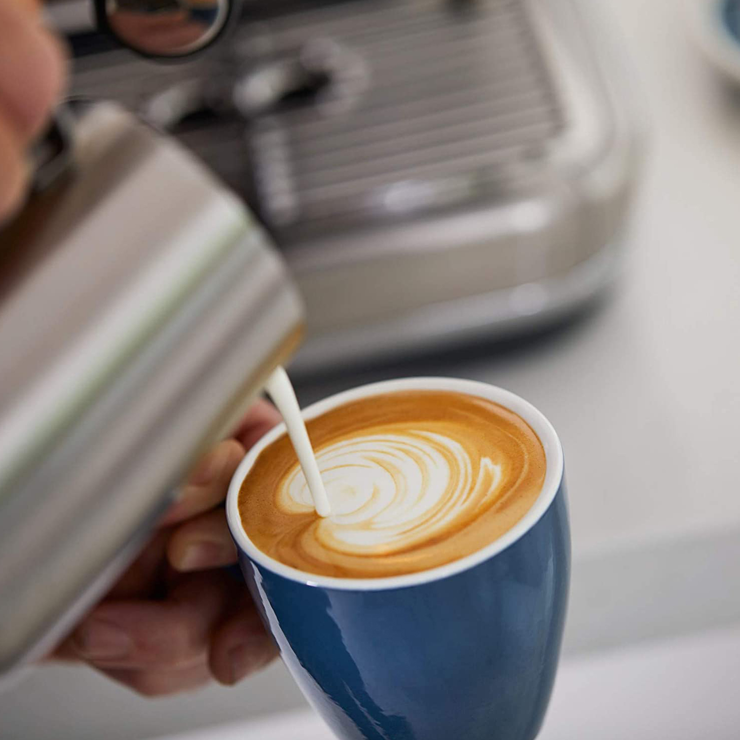 Una mano versa latte in una tazza di caffè blu, creando un motivo latte art. Una macchina per il caffè è sullo sfondo.