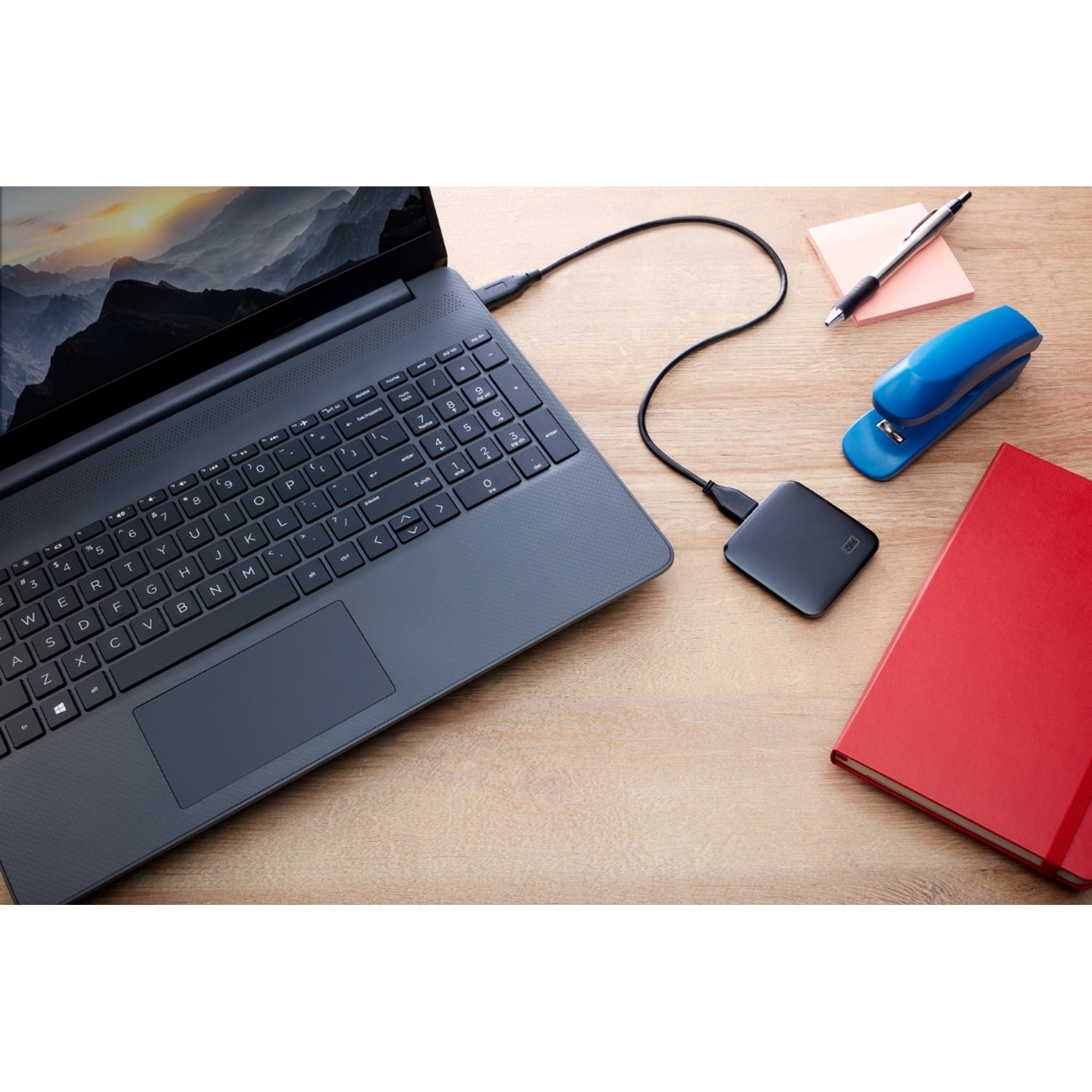 Laptop conectado a un disco duro externo. Un bolígrafo, grapadora y cuaderno están sobre el escritorio.