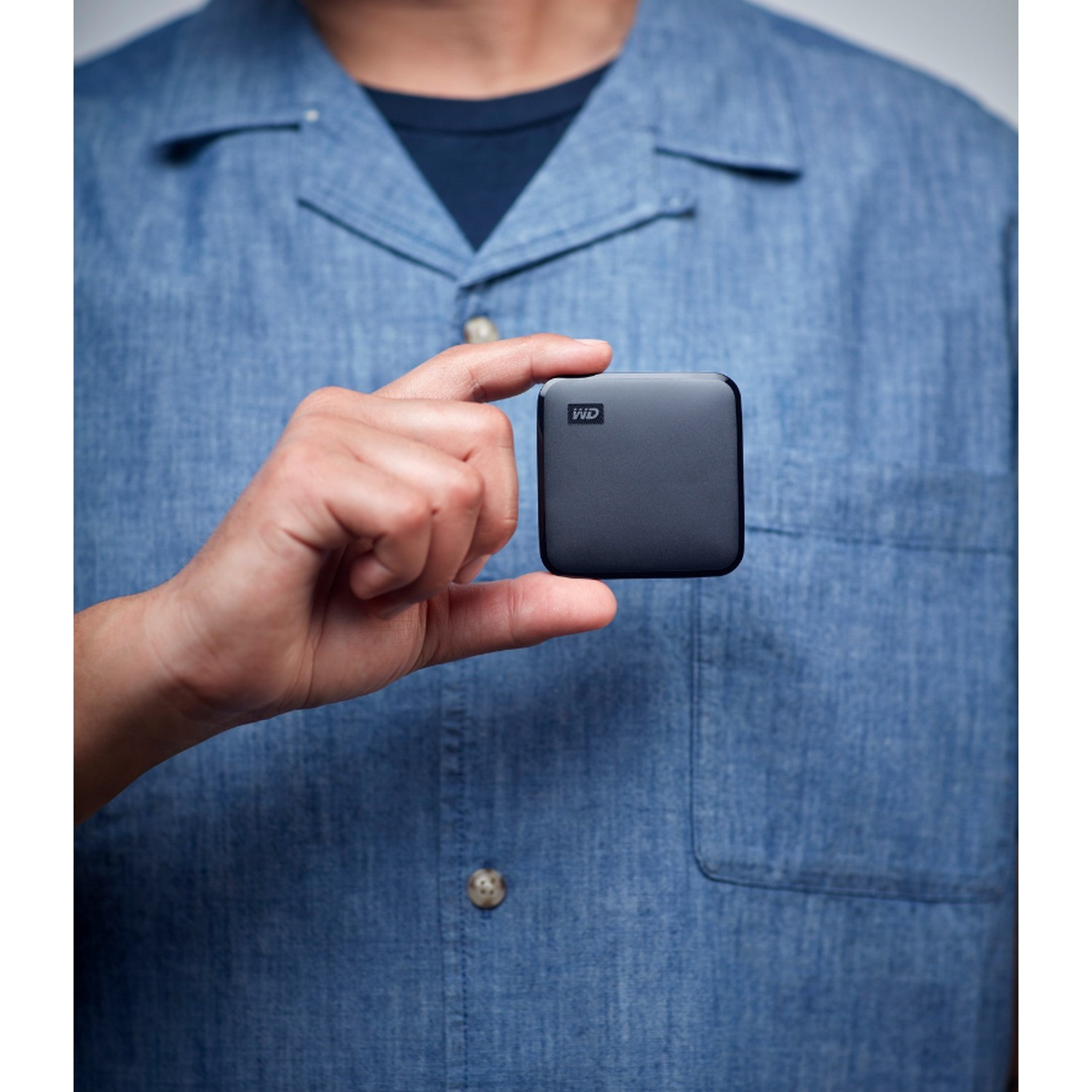 Una mano sostiene un disco duro externo pequeño y negro con un logotipo de WD. La persona lleva una camisa azul.