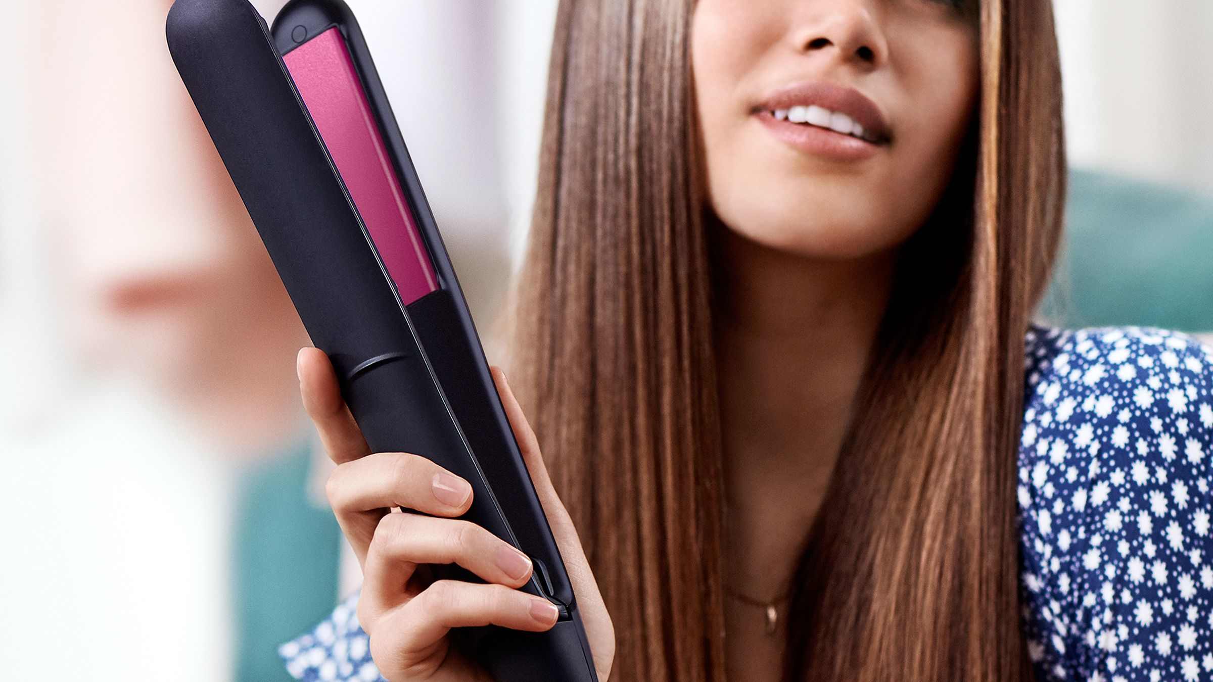 Una mujer sosteniendo una plancha de pelo negra y rosa, con cabello castaño largo en el fondo.