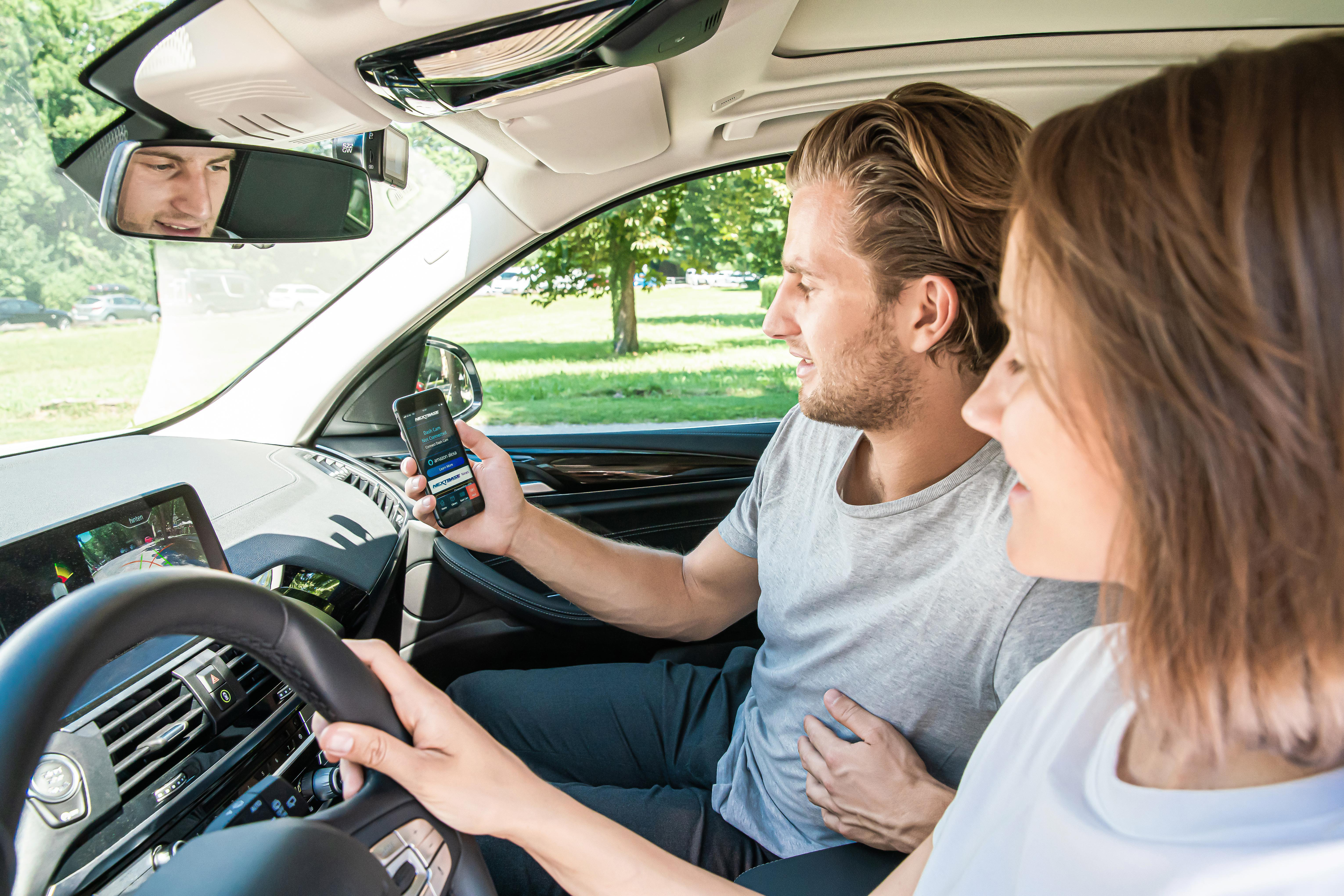 Ein Paar im Auto, Mann hält Telefon mit App, Frau fährt, Bäume im Hintergrund.