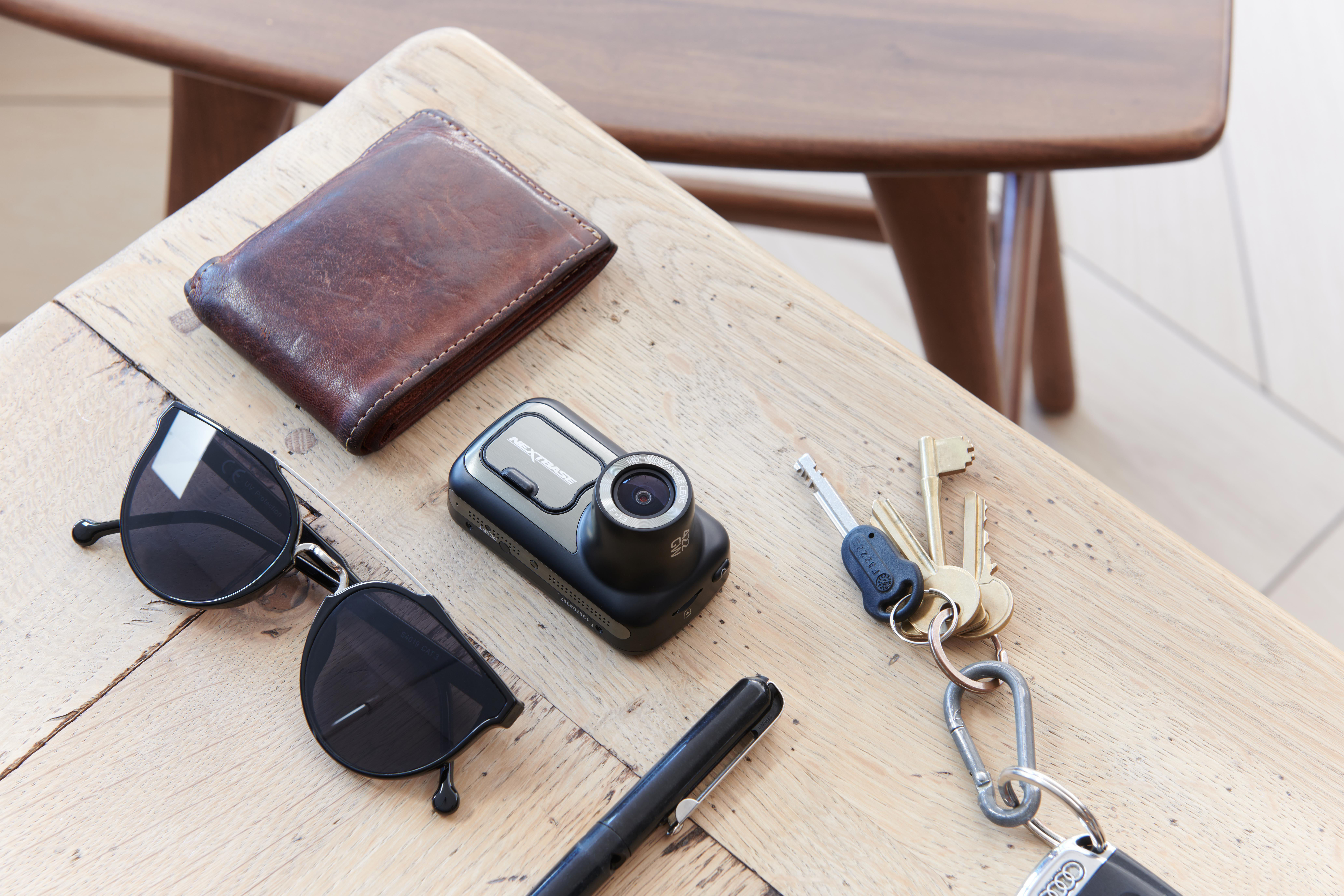 Ein Tisch mit braunem Portemonnaie, Kamera, Sonnenbrille, Stift, Schlüsseln und Stuhl im Hintergrund.