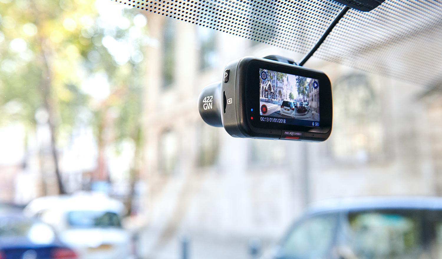 Eine schwarze Dashcam ist an einer Windschutzscheibe montiert und zeigt ein Auto und Gebäude auf dem Bildschirm.