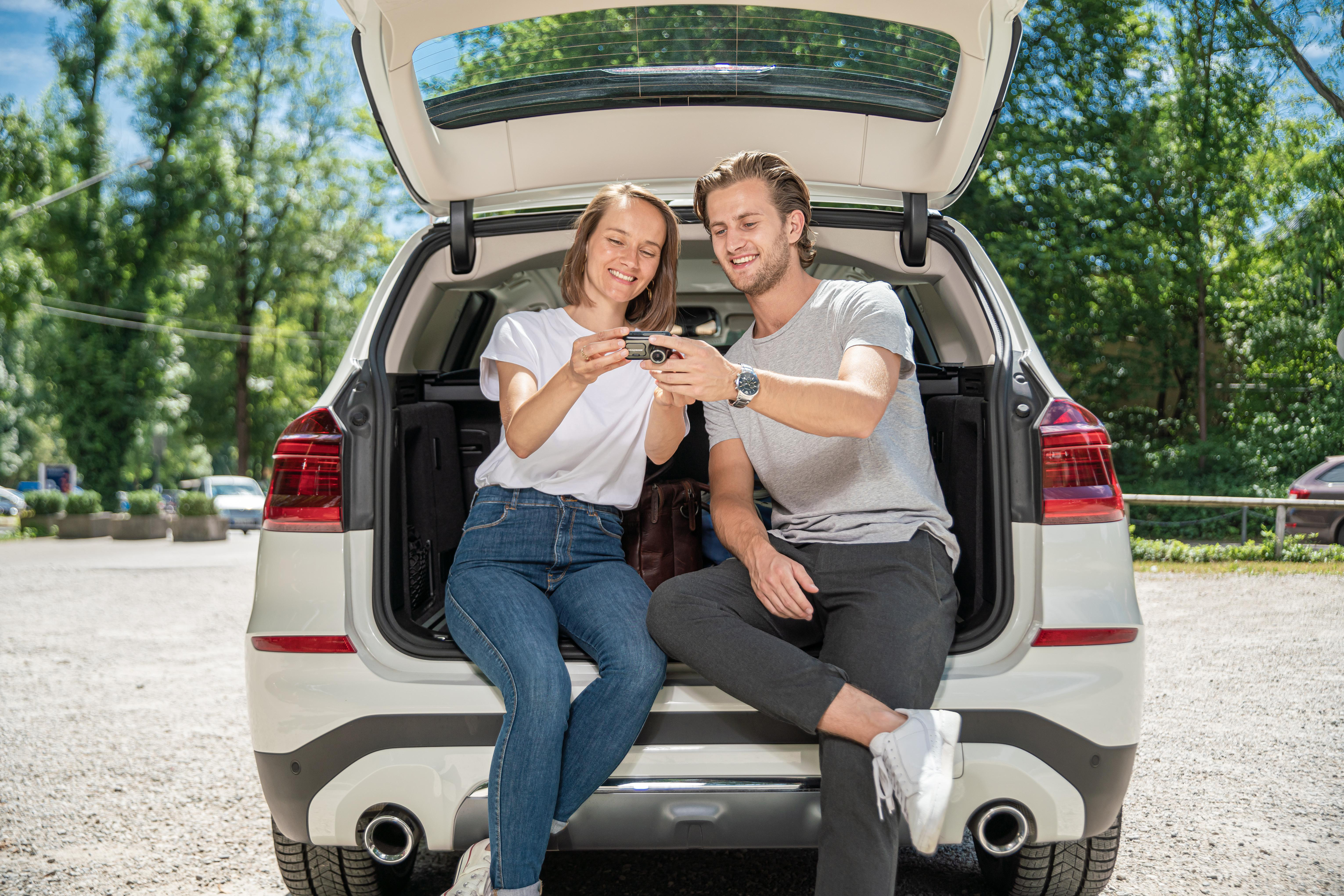 Ein Paar sitzt in einem offenen Kofferraum und betrachtet eine Kamera. Bäume im Hintergrund.