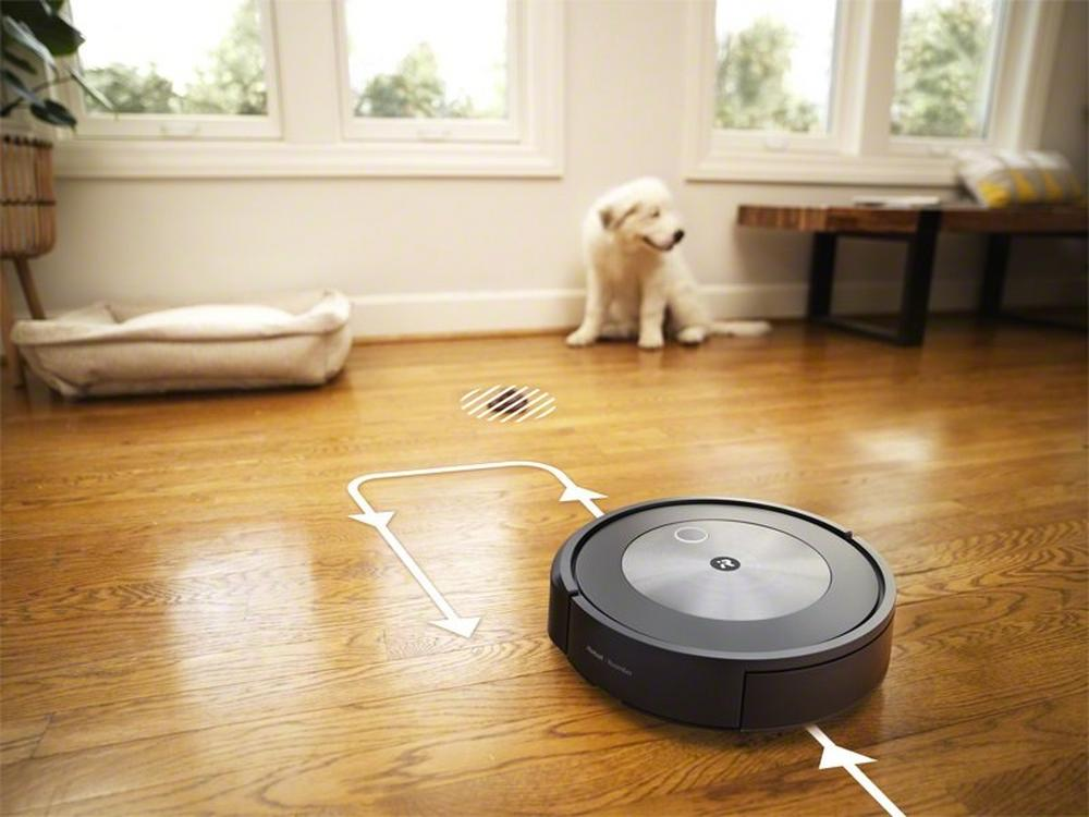 Robotstofzuiger op houten vloer aan het schoonmaken. Een hond zit op de achtergrond. Ramen boven.