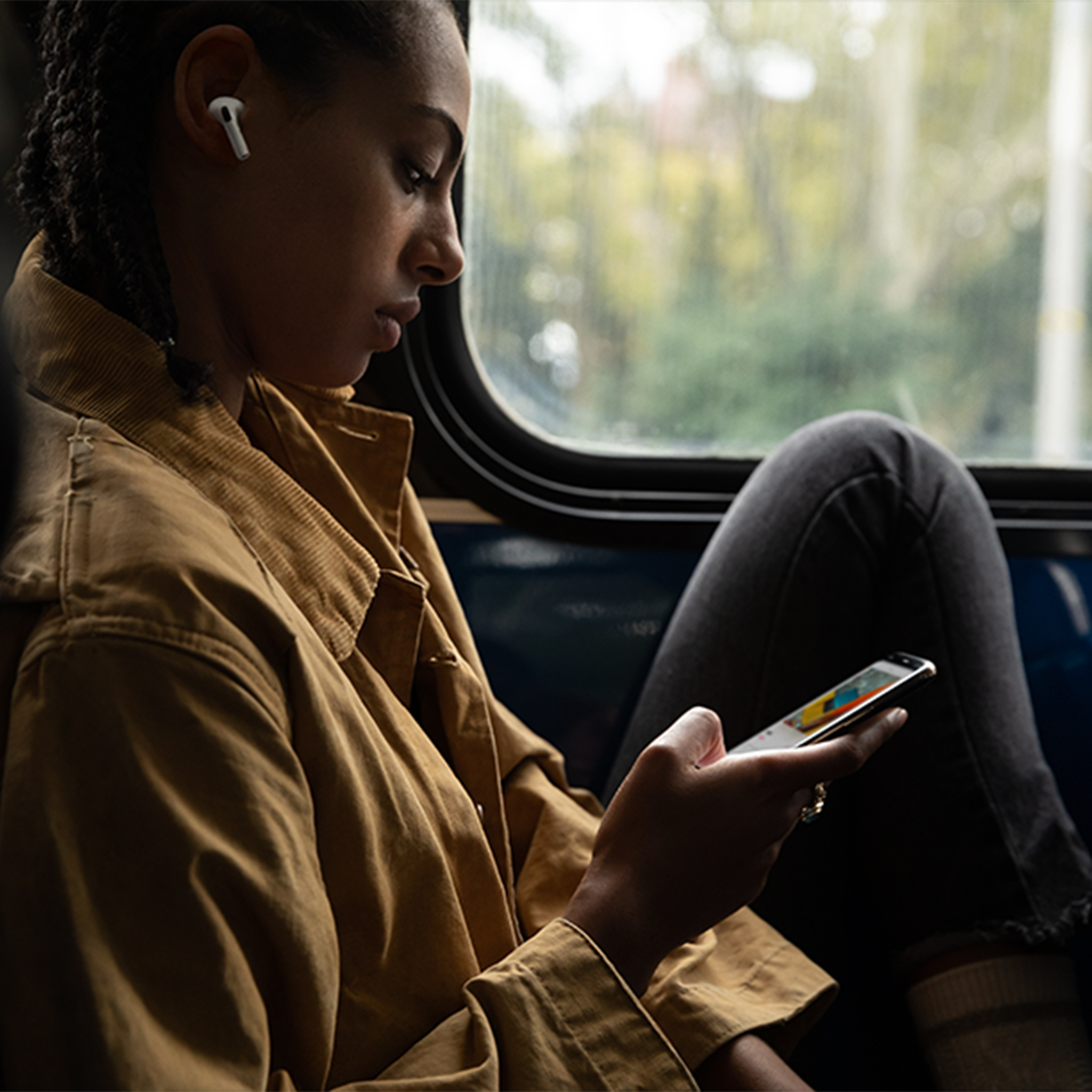 Eine Frau sitzt in einem Bus, trägt einen Ohrhörer und schaut auf ihr Handy. Sie trägt eine braune Jacke und dunkle Hosen.
