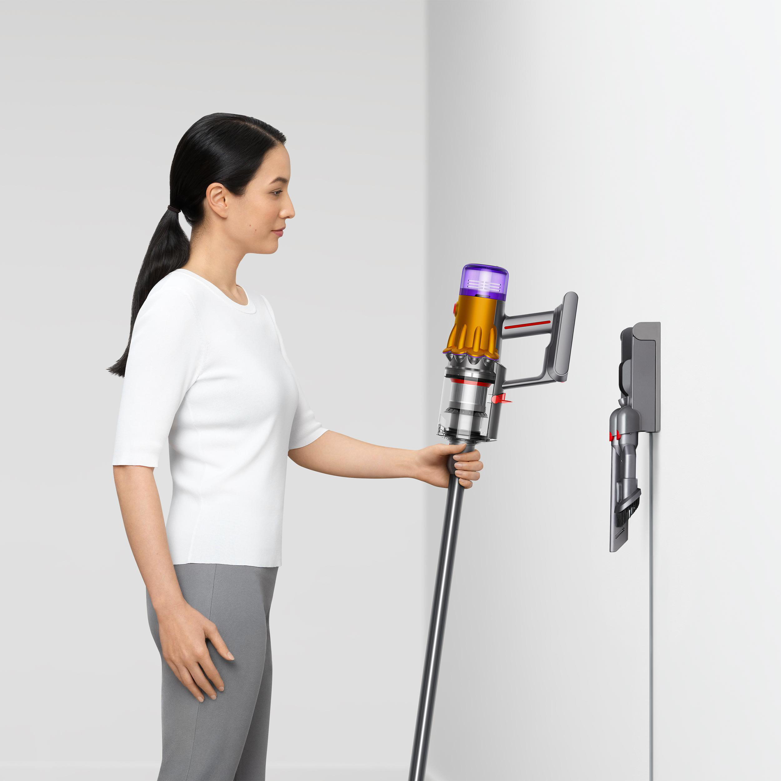 Frau hält einen Dyson-Staubsauger. Rechts ist ein an der Wand montiertes Ladegerät. Weißer Hintergrund.