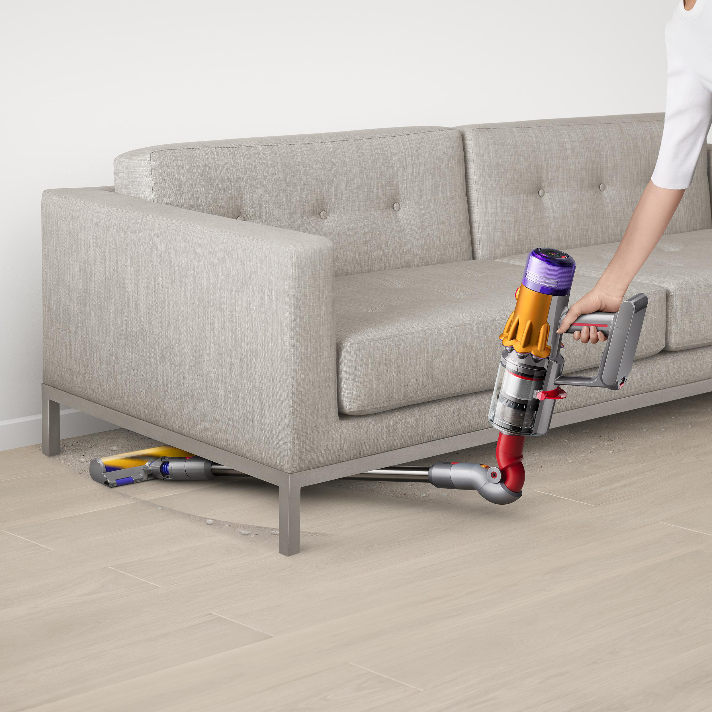 Eine Person saugt mit einem gelb-violetten Dyson-Staubsauger unter einem grauen Sofa auf hellem Holzboden.