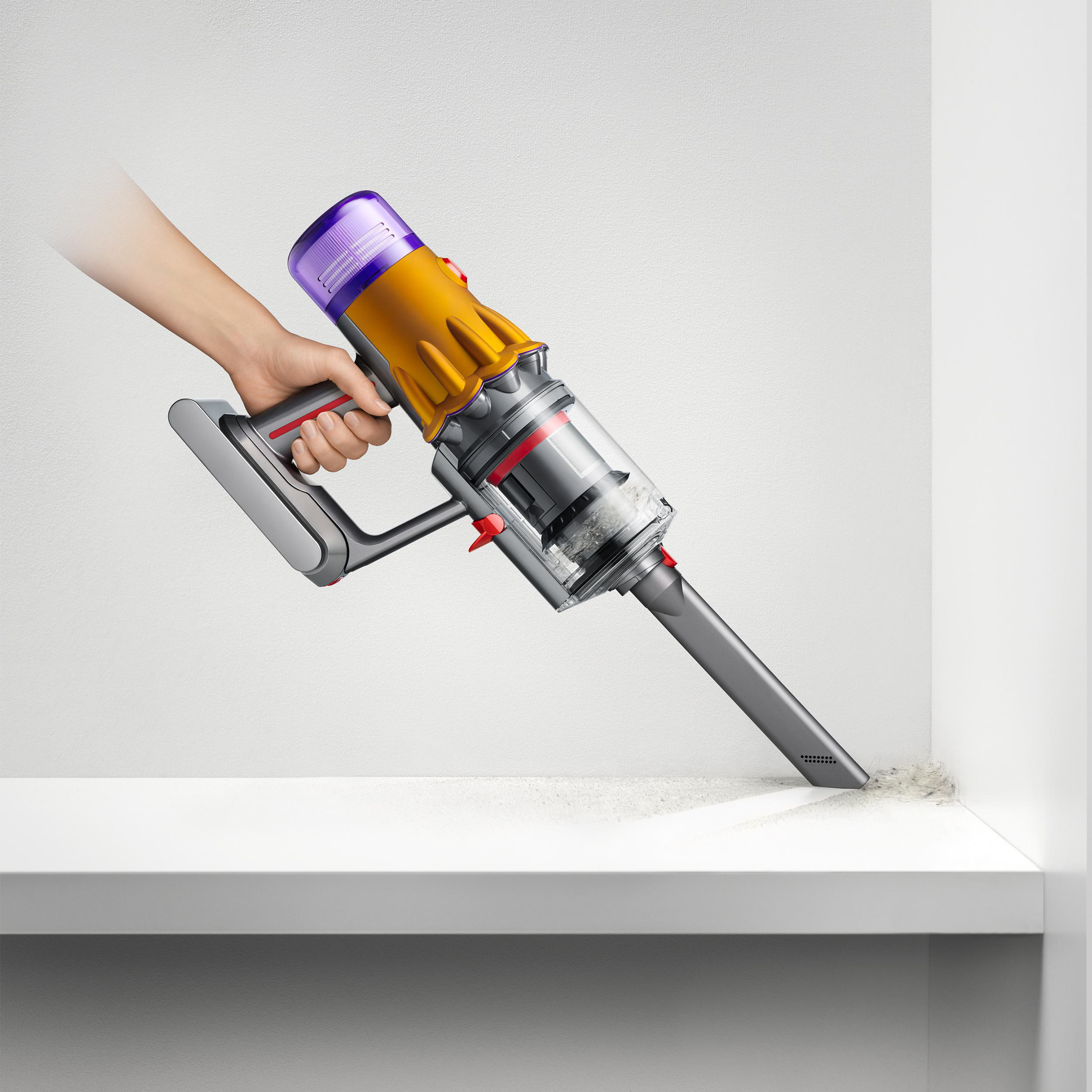 Hand hält gelb-violetten Staubsauger, der eine Ecke reinigt. Weißer Hintergrund und Oberfläche.