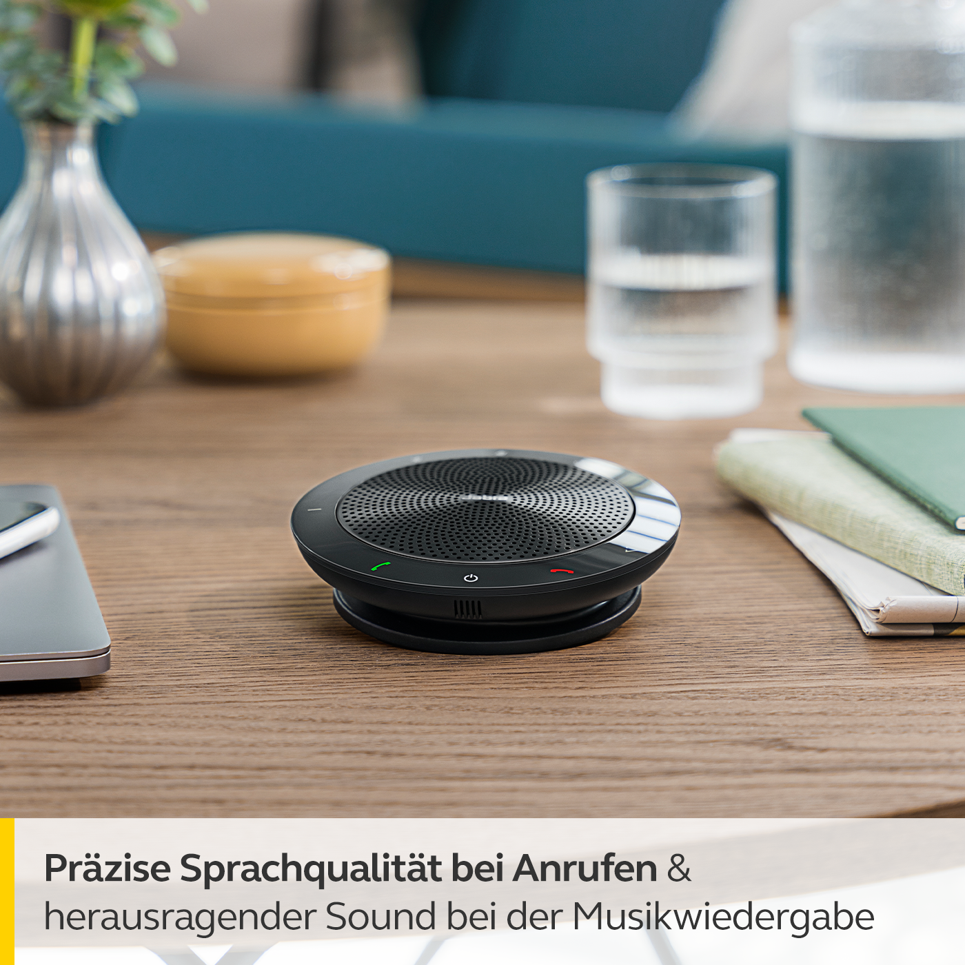 Ein schwarzer Lautsprecher auf einem Holztisch mit Laptop, Wasserglas und anderen Gegenständen.