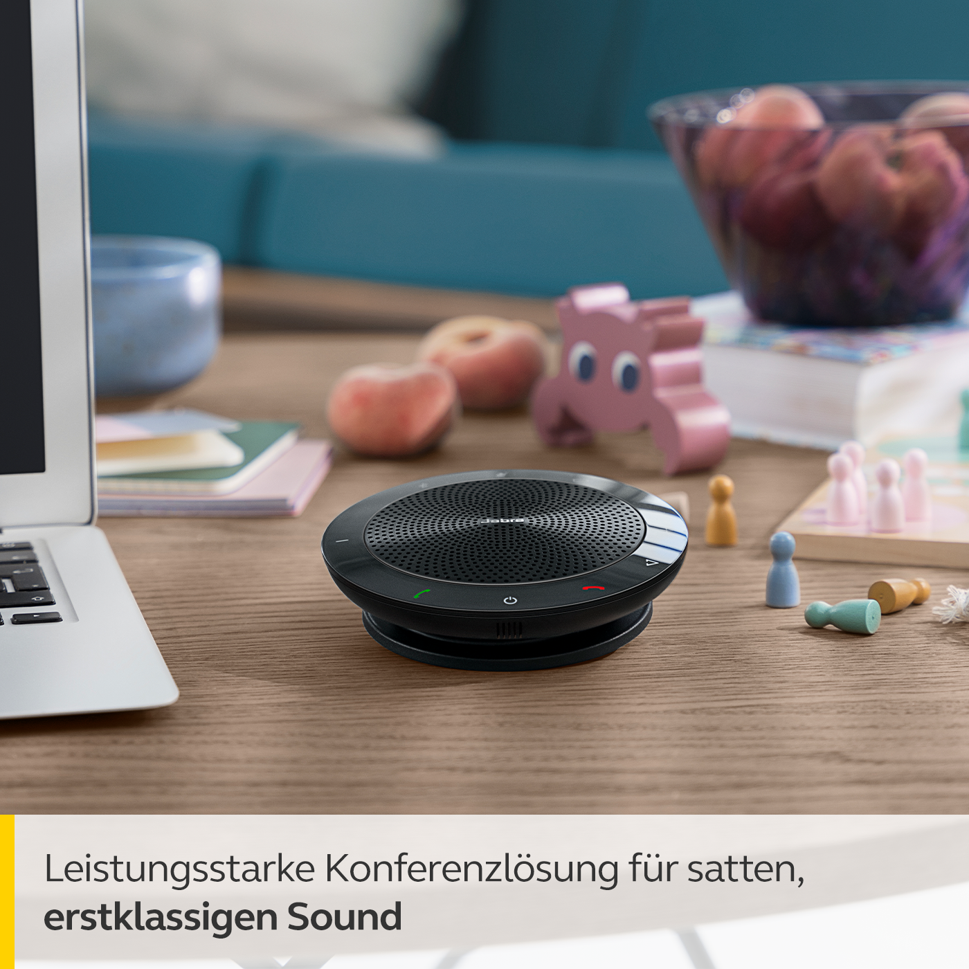 Schwarze Jabra Freisprecheinrichtung auf Holzschreibtisch. Weitere Gegenstände: Laptop, Obstschale und Figuren.