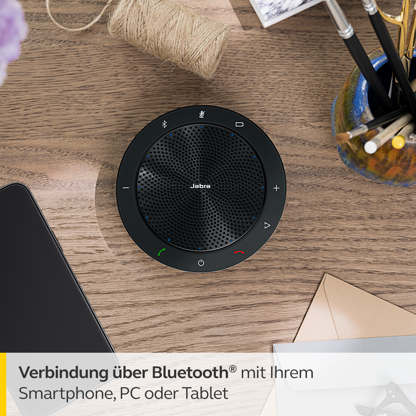 Schwarzes Jabra-Freisprechgerät auf einem Holztisch. Telefon, Stifte und Notizblock sichtbar.