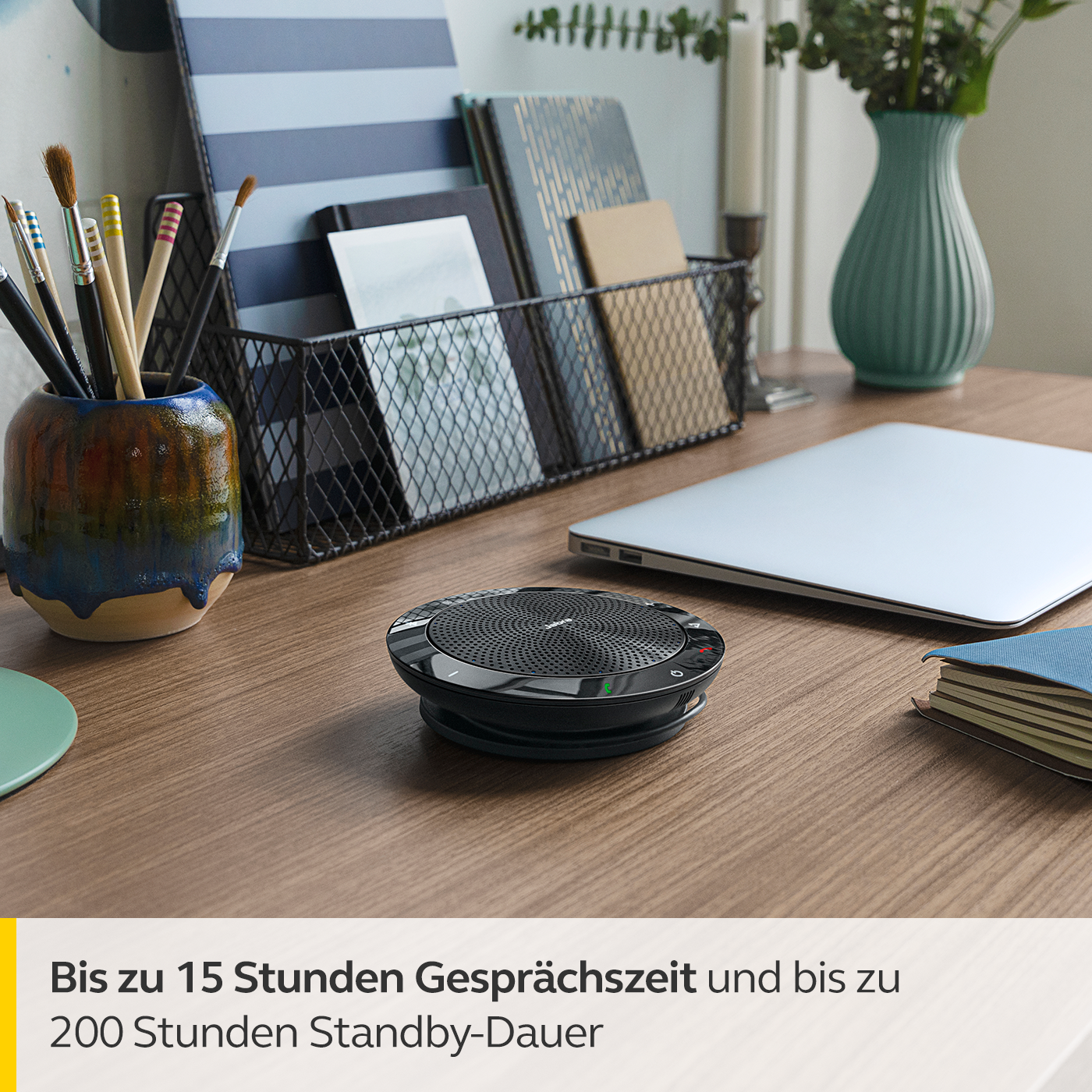 Ein schwarzer Lautsprecher auf einem Schreibtisch mit Laptop, Notizbüchern und Kunstzubehör.