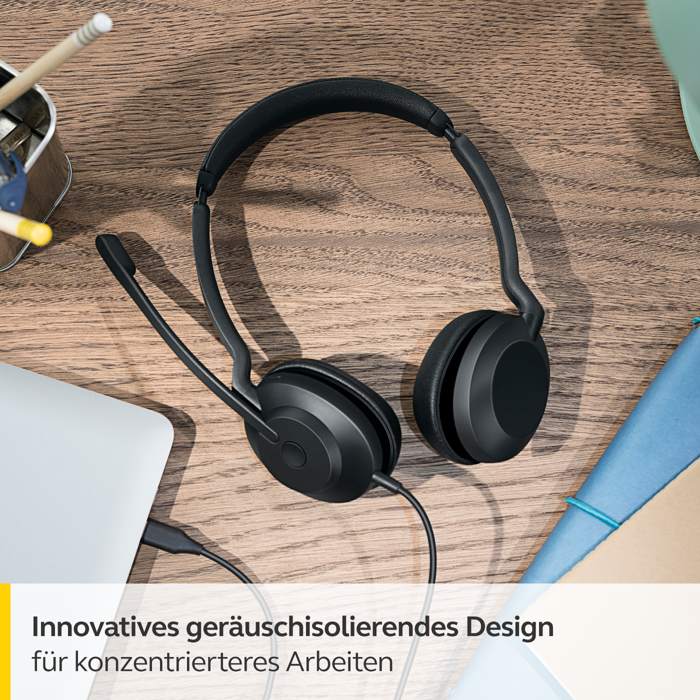 Ein schwarzes Jabra-Headset auf einem Holzschreibtisch, mit einem Laptop und einem blauen Notizbuch in der Nähe.
