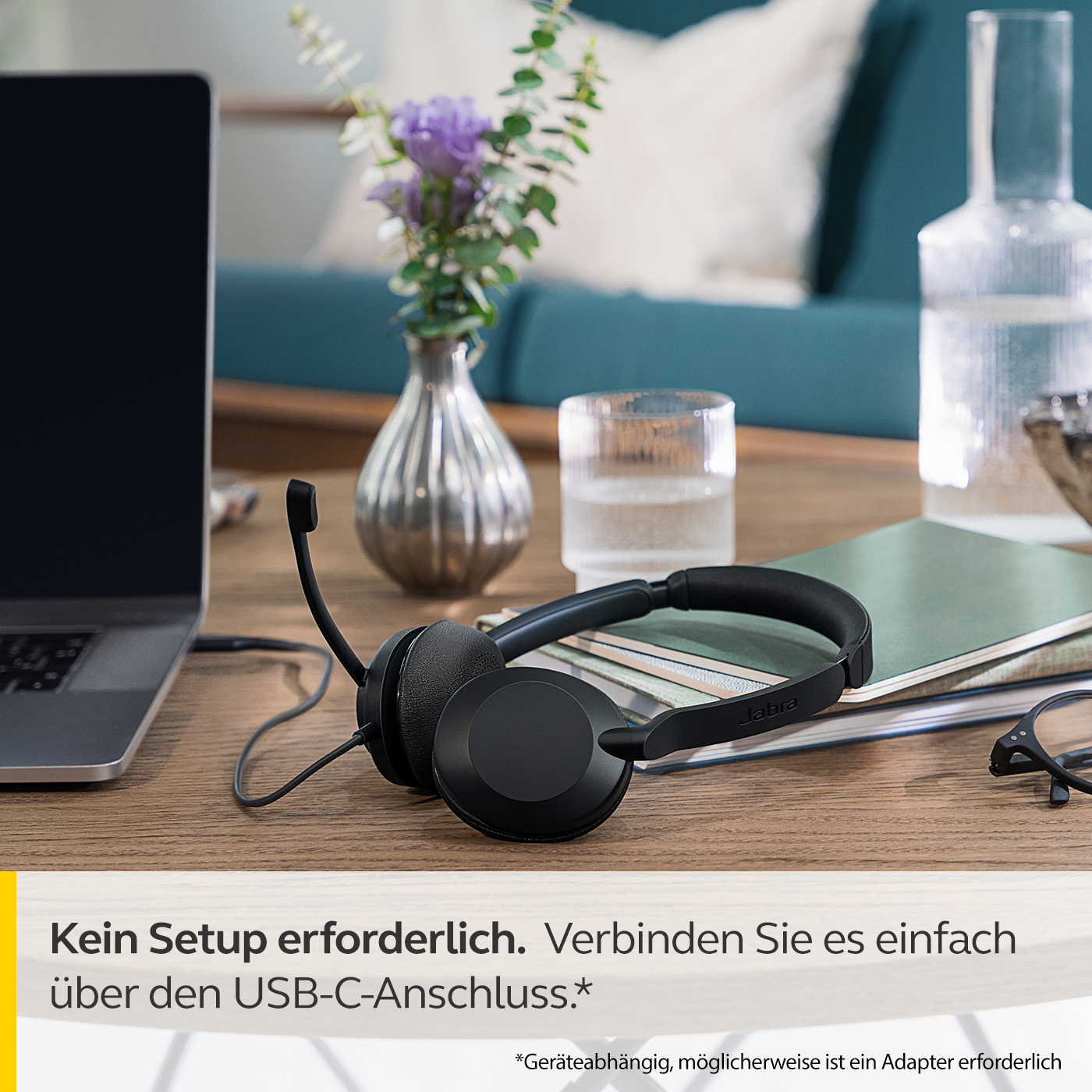 Schwarzes Headset mit flexiblem Mikrofon, das auf einem Holztisch liegt. Ein Glas Wasser und ein Laptop sind ebenfalls auf dem Tisch.