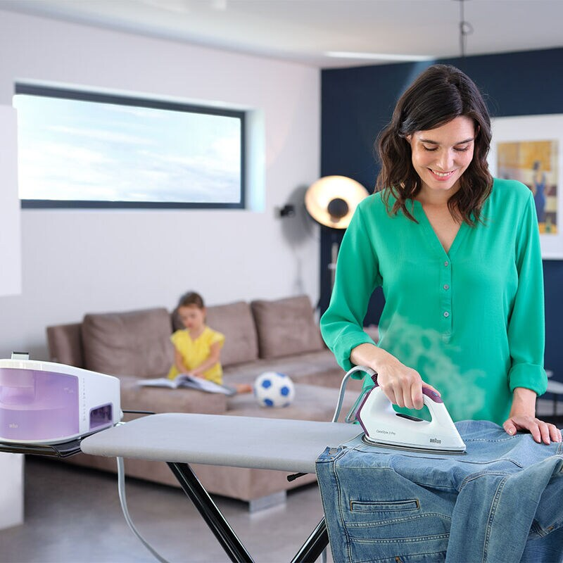 A woman ironing jeans on an ironing board. A child and a sofa are in the background.