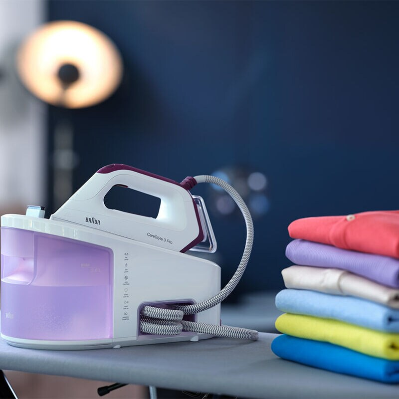 An iron and ironing board with folded clothes. A blue wall and a lamp are in the background.