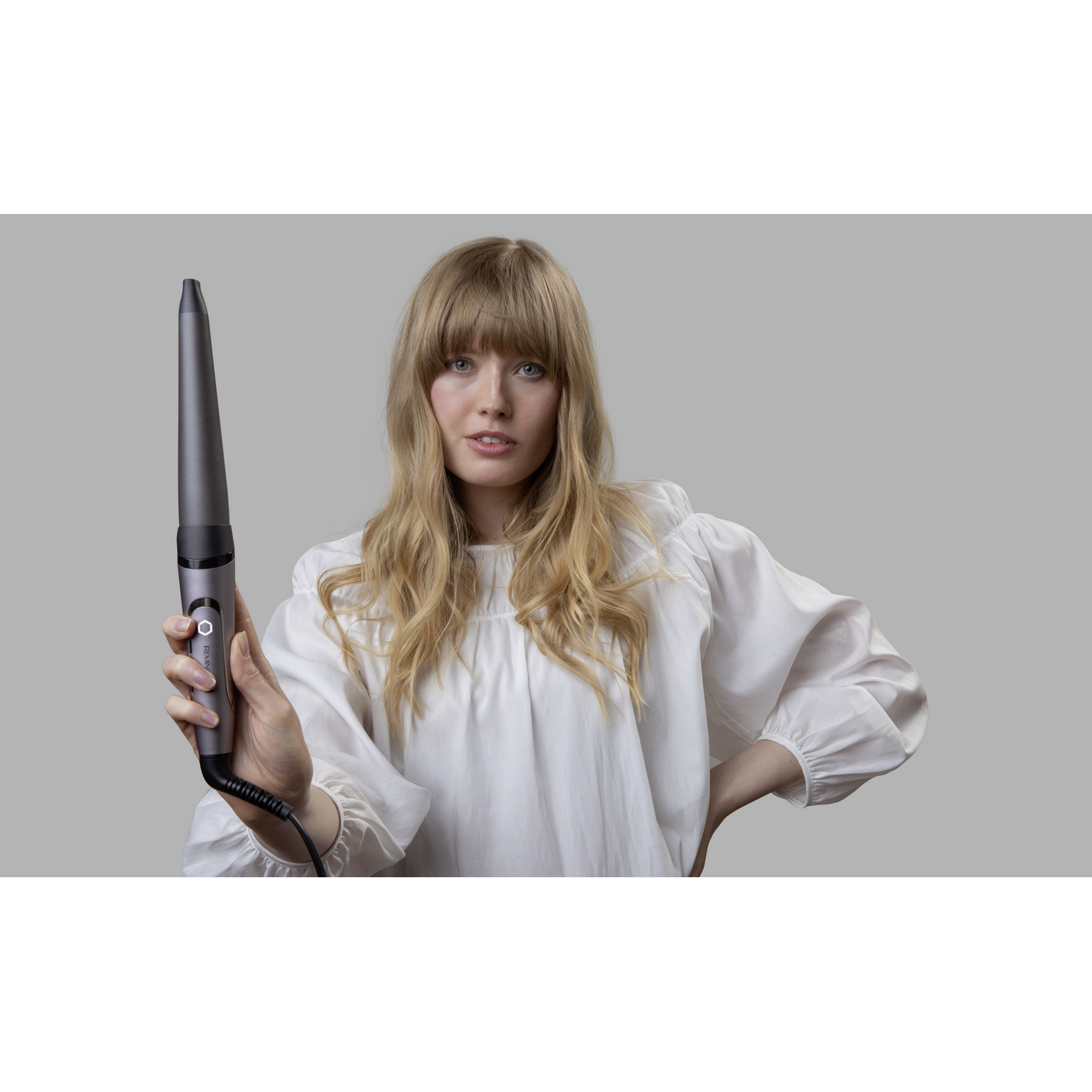 Mujer rubia sostiene un rizador, mirando a la cámara. Lleva una blusa blanca.