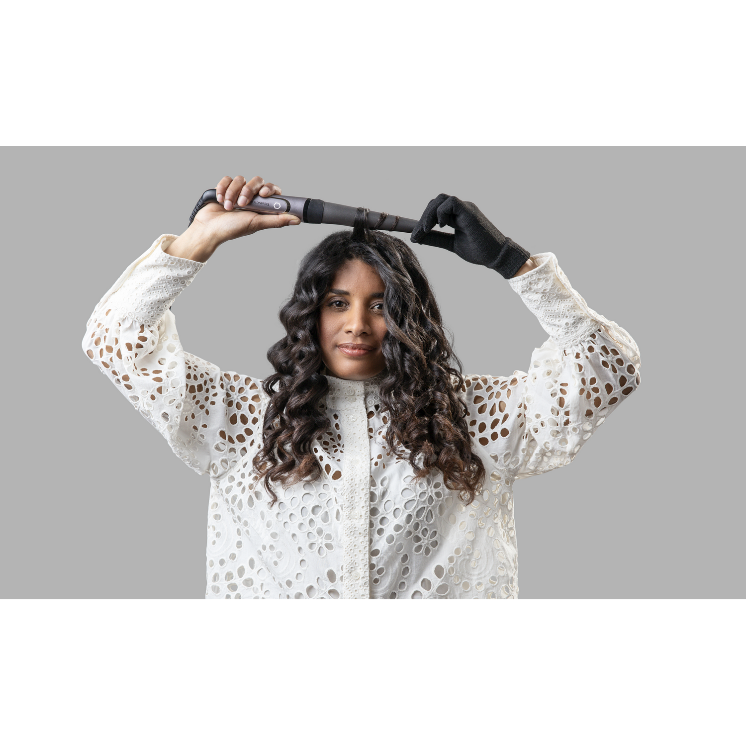 Mujer con cabello rizado usando rizador, camisa blanca, guantes negros.