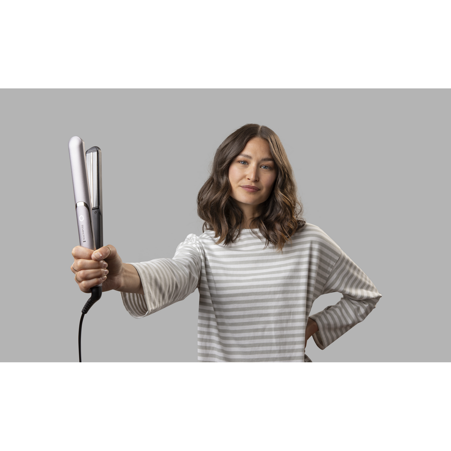 Una mujer sosteniendo una plancha para el pelo, con el pelo peinado, sobre un fondo gris.