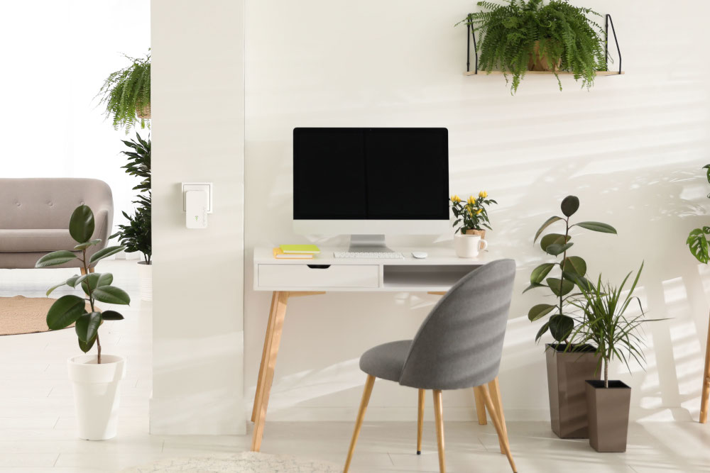 Een bureau met een computer en planten, met een wit apparaat aan de muur op de achtergrond.