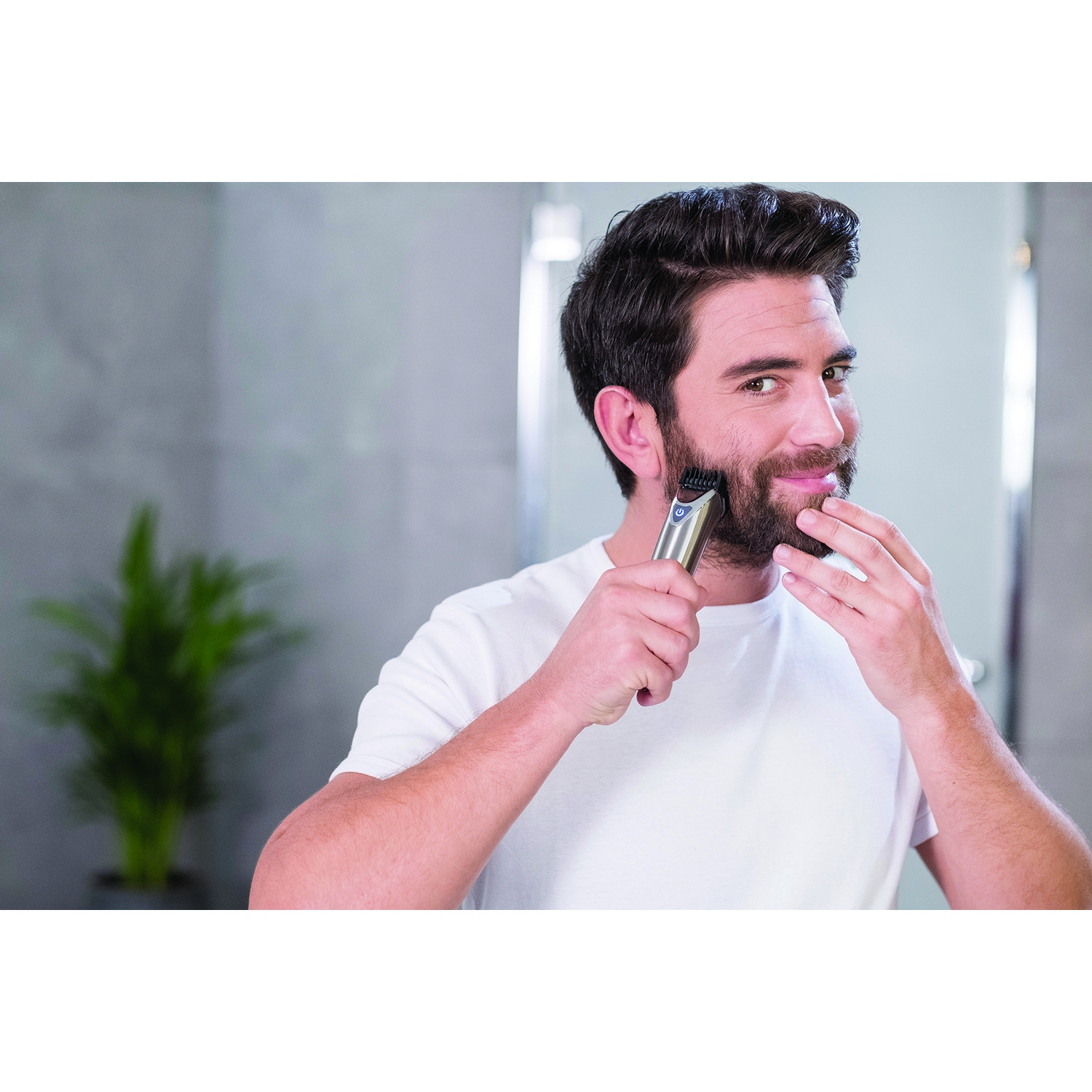 Un hombre se afeita la barba con una recortadora eléctrica plateada frente a un espejo en un baño.
