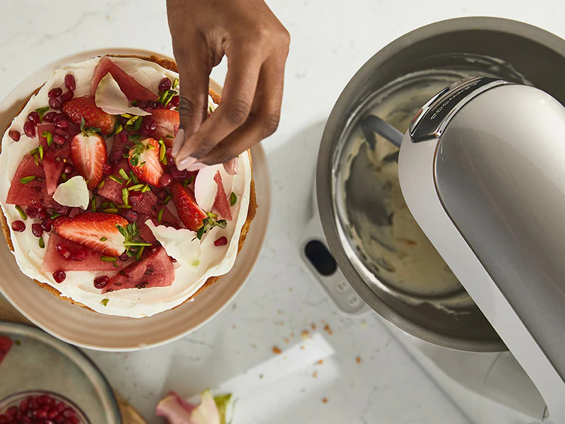 Cake being decorated with fruit. A hand adds strawberries and pistachios.