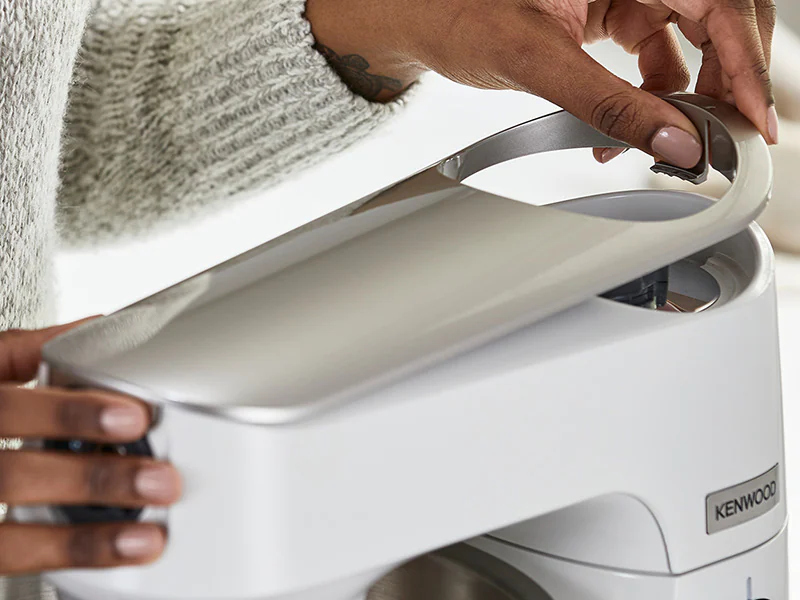 Hands opening the lid of a white Kenwood kitchen appliance.