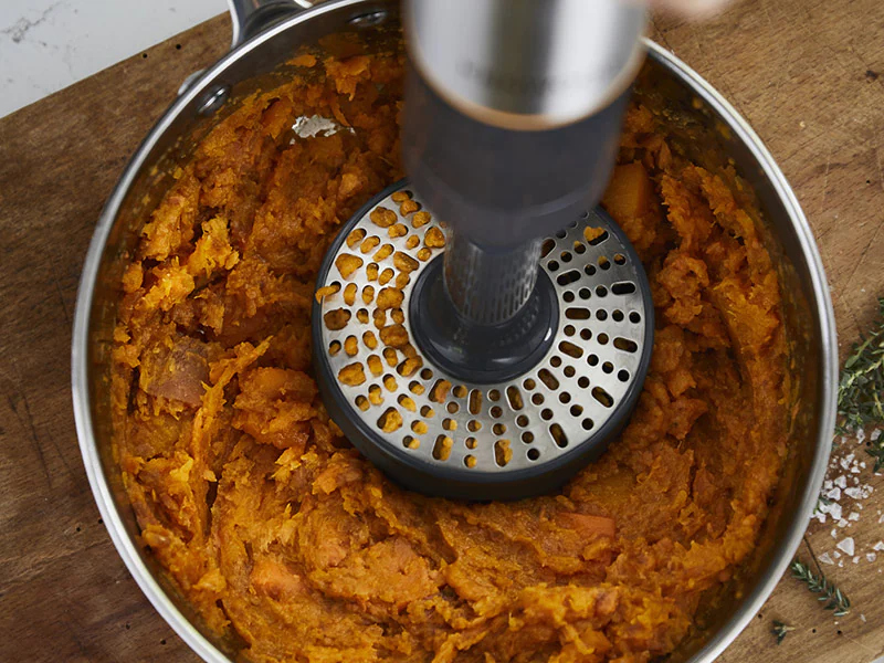 Hand blender in a pot of mashed sweet potatoes. The pot is on a wooden surface.