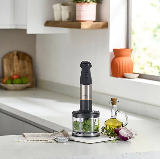 Hand blender in a glass, ingredients on the counter with oil and onion.