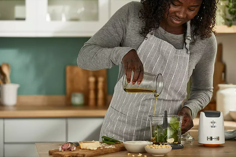 Femme versant de l'huile dans un mixeur avec du basilic et des pignons de pin. Scène de cuisine, éclairage vif.
