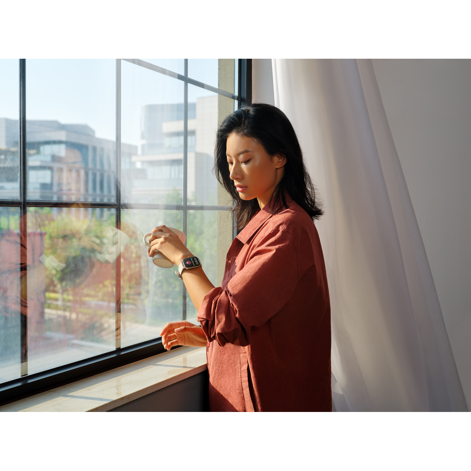 Mujer mirando por la ventana, taza en mano, camisa naranja, reloj inteligente. Luz del día brillante.