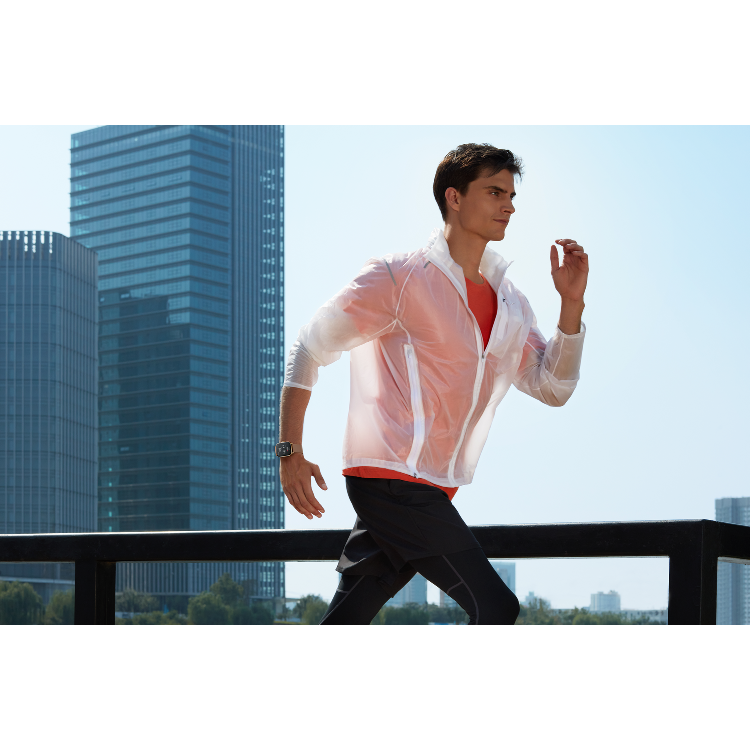 Hombre corriendo al aire libre, con una chaqueta y un reloj. Edificios y cielo azul al fondo.