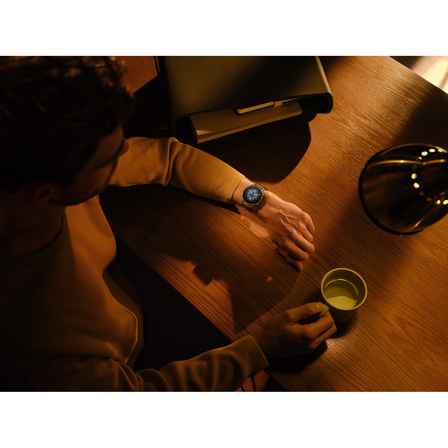 Hombre en escritorio, reloj, taza. Lámpara visible. Fondo oscuro.