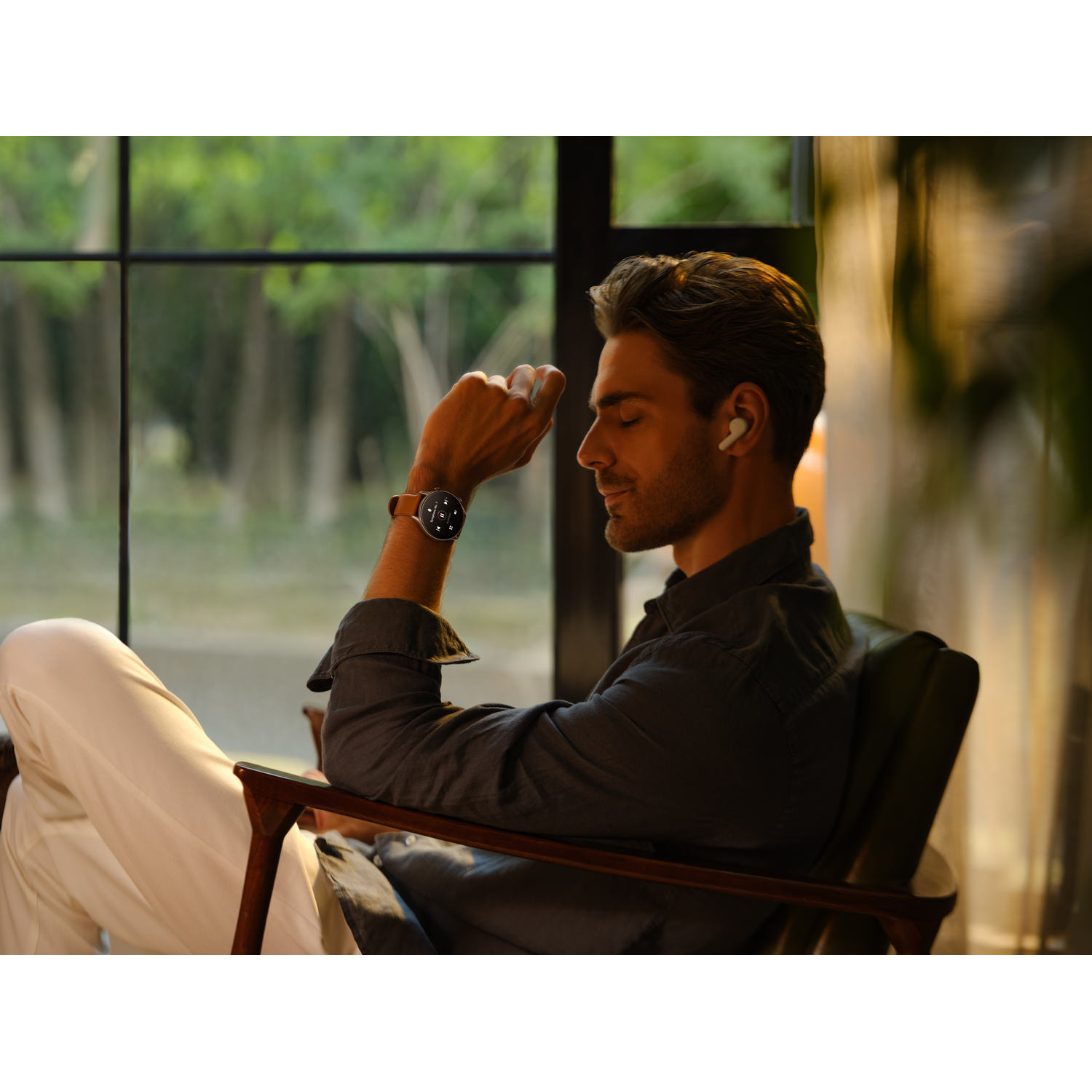 Hombre con reloj y auriculares, sentado en una silla. Una ventana está en el fondo.