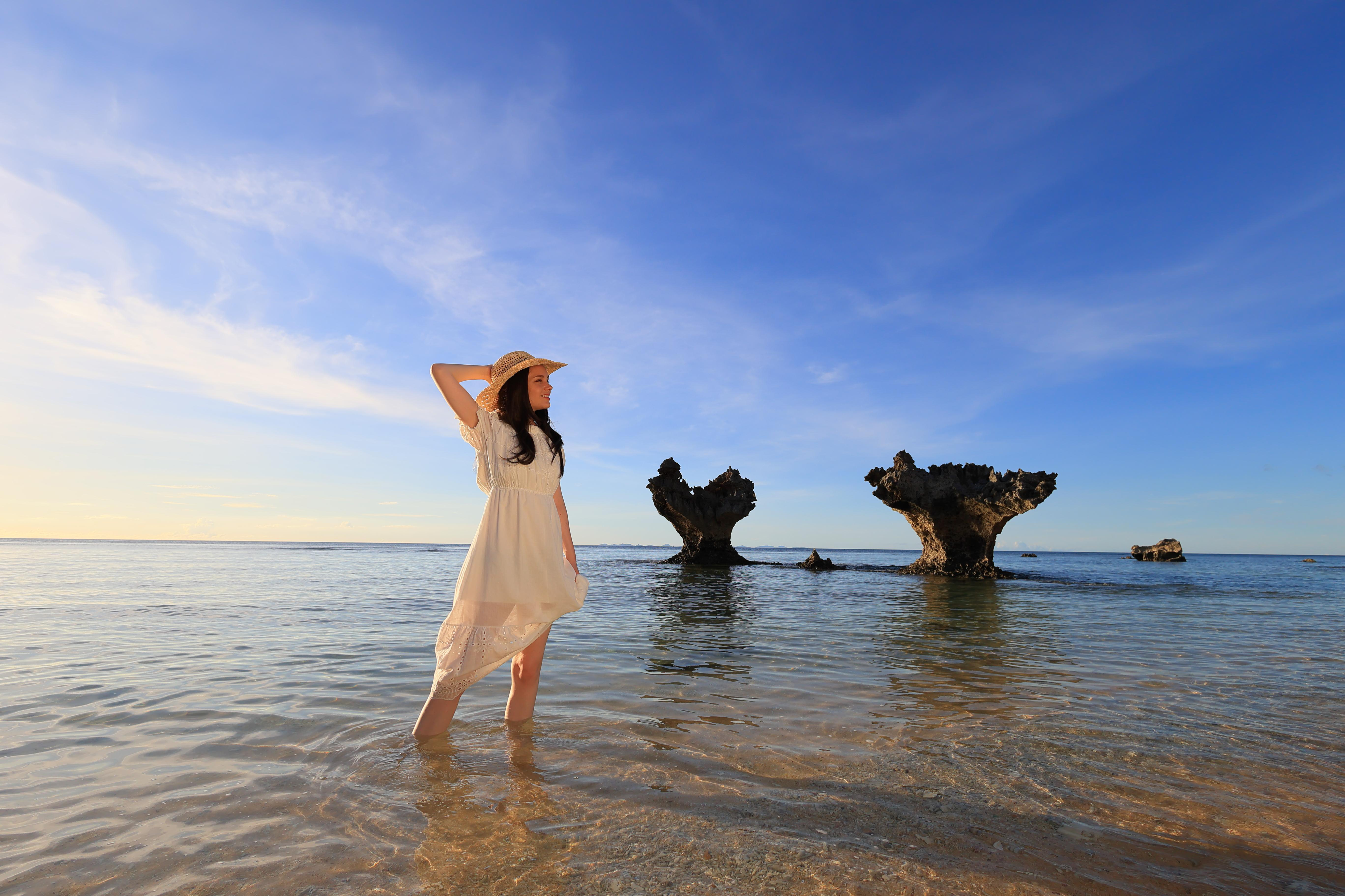 Frau in weißem Kleid und Hut steht im flachen Wasser am Strand. Klarer blauer Himmel.