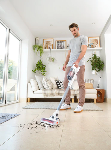 Homme utilisant un aspirateur sur un sol sale. La pièce a des plantes, un canapé et des cadres.