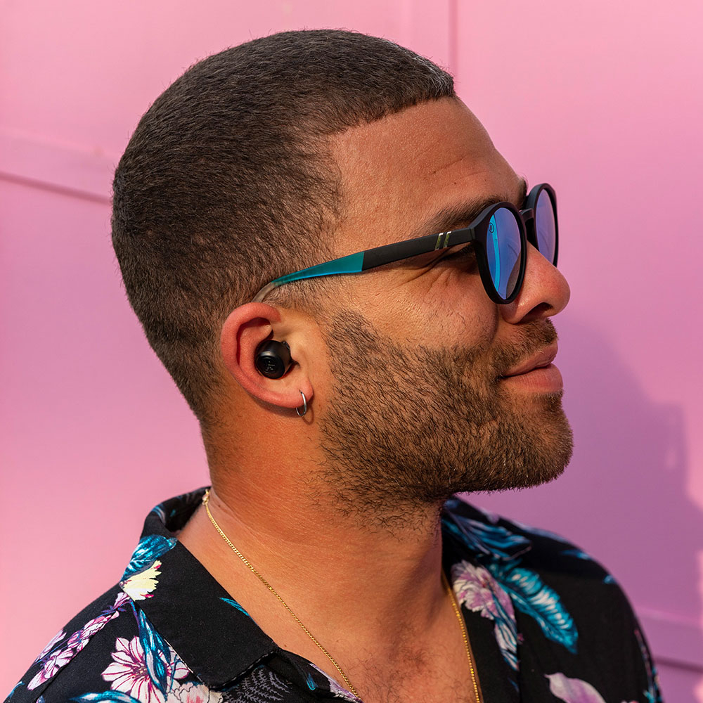 Homme portant des lunettes de soleil et des écouteurs, fond rose, regardant ailleurs.
