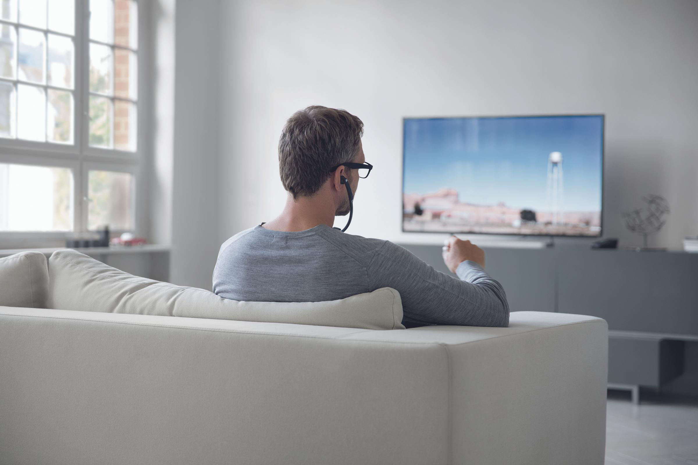 Mann mit Brille und Ohrhörern sitzt auf Sofa und schaut Fernsehen. Einfaches Wohnzimmer.