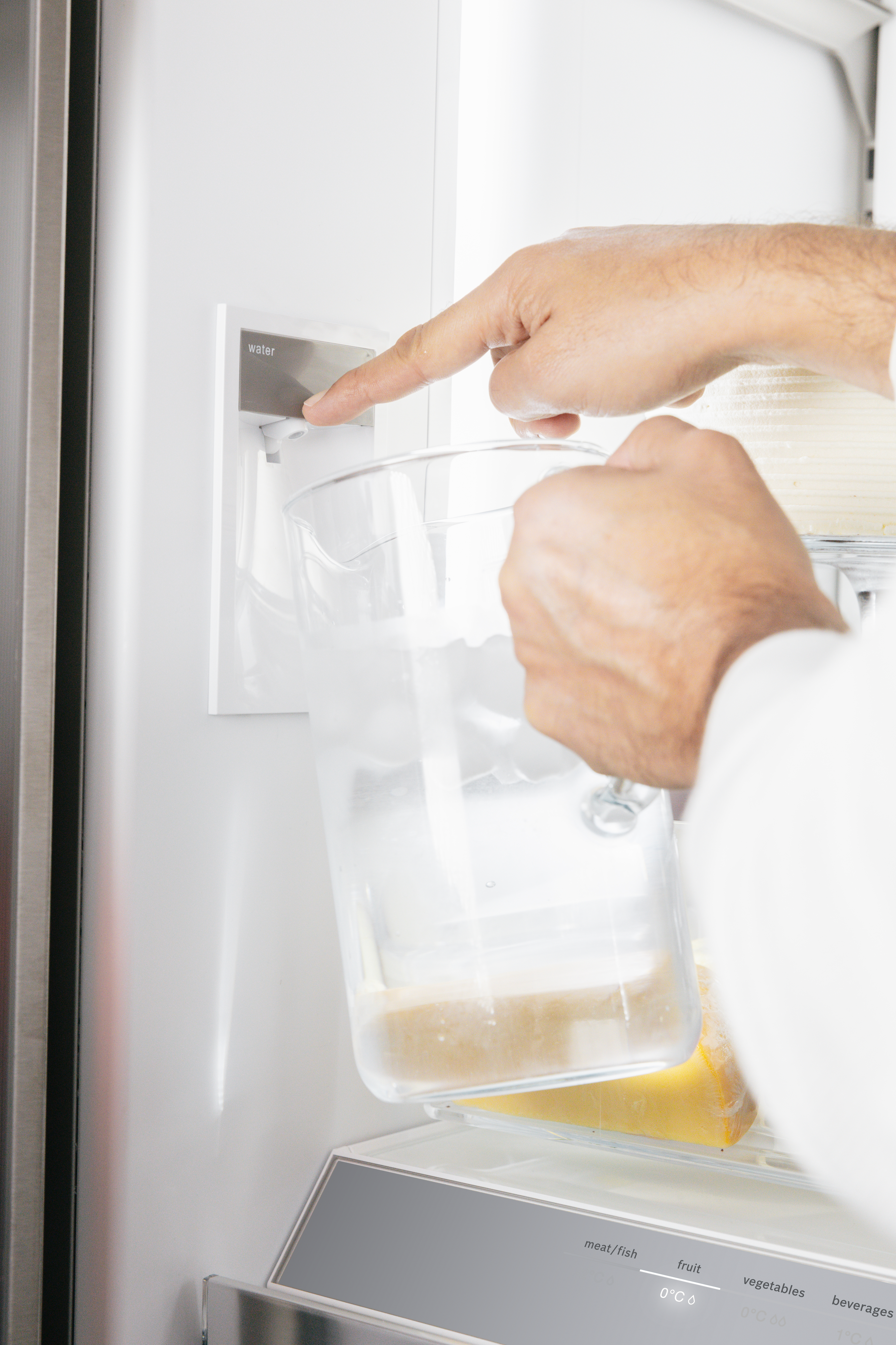 Eine Hand drückt eine Wassertaste, um einen transparenten Krug in einem Kühlschrank zu füllen.