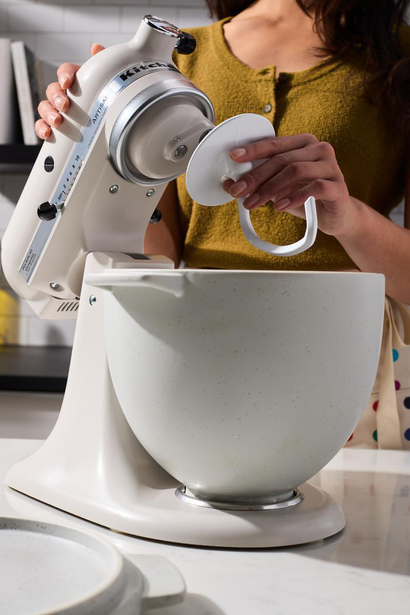 Eine Frau benutzt einen KitchenAid-Küchenmixer und hält einen Aufsatz fest.