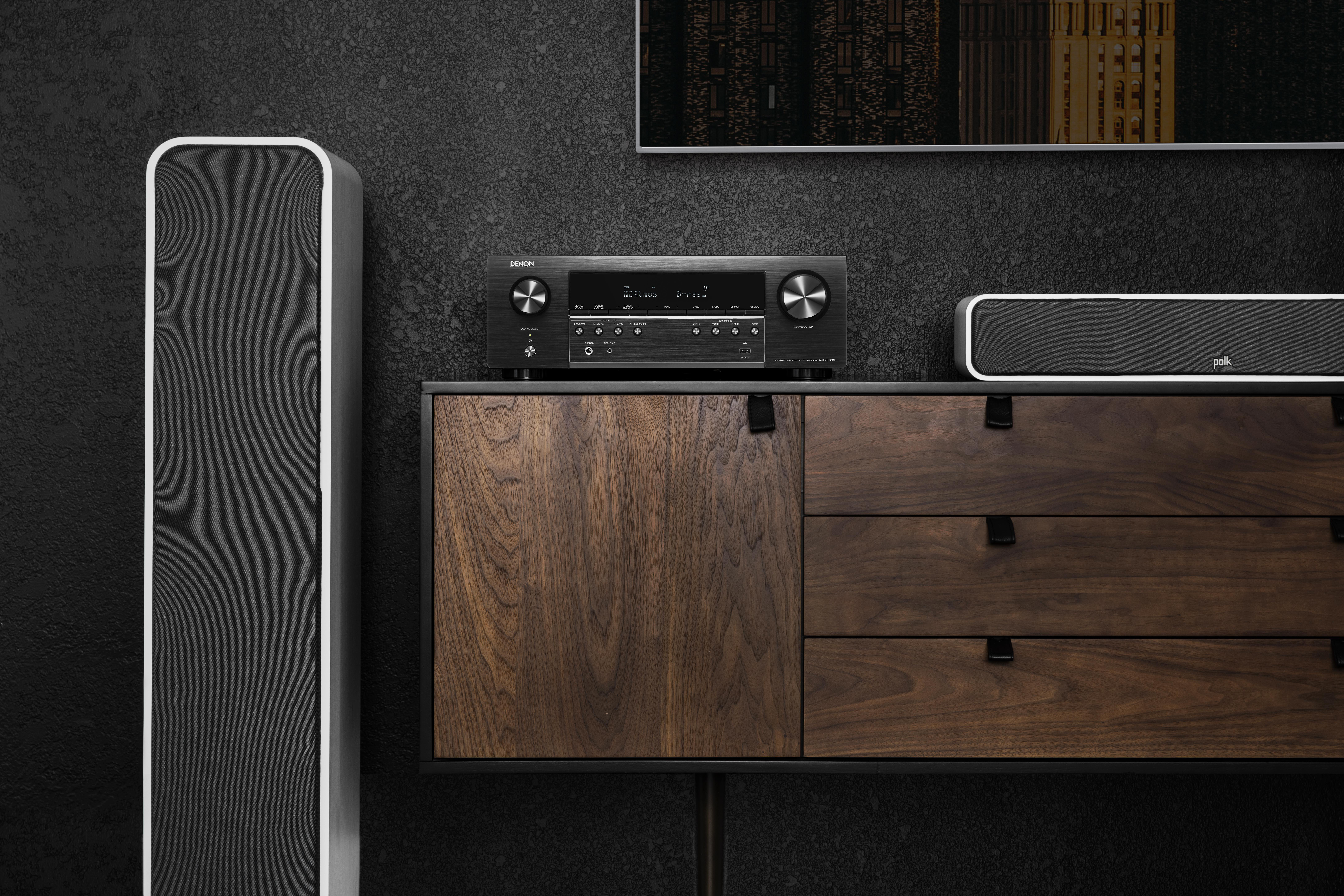 Denon-Receiver auf einem Holzschrank, flankiert von Lautsprechern und einem TV an dunkler Wand.