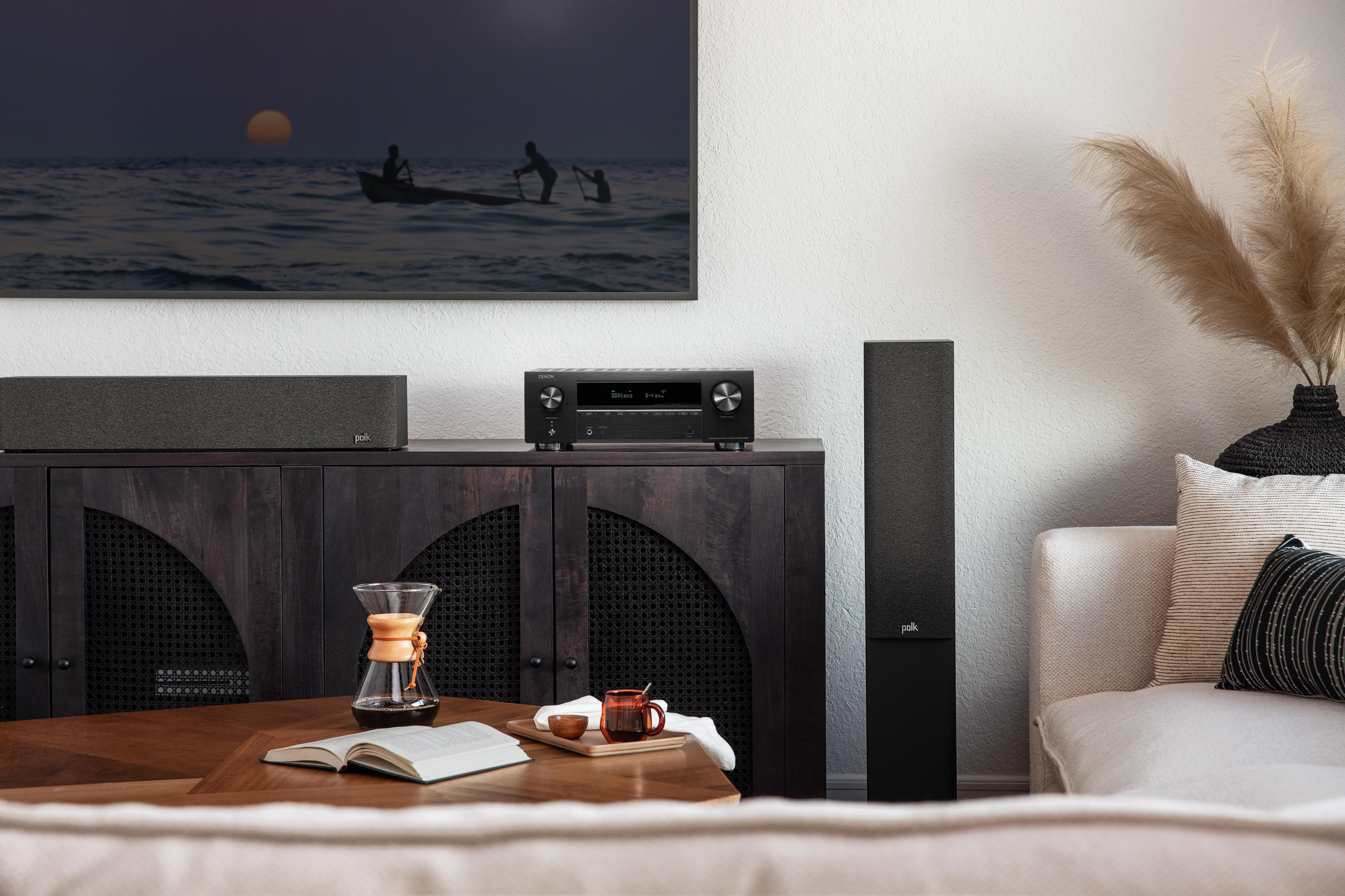 Wohnzimmer mit Fernseher, Soundbar und Lautsprechern auf einem dunklen Schrank. Ein Couchtisch mit Buch und Kaffee.