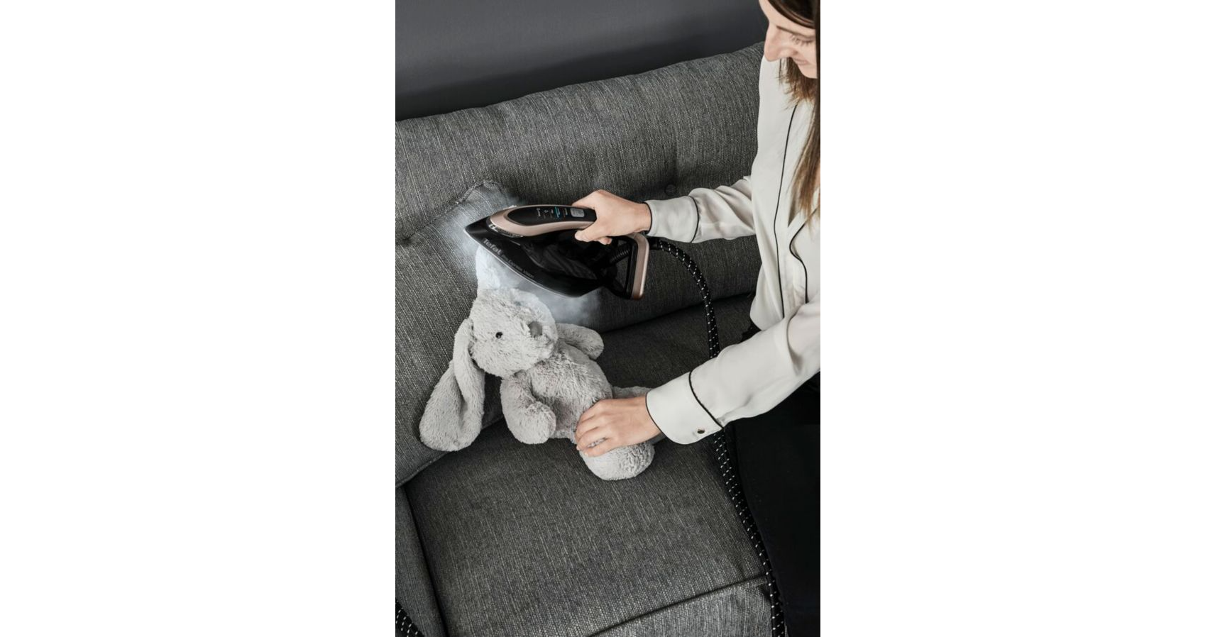 A woman steaming a gray stuffed bunny on a sofa with a black iron.